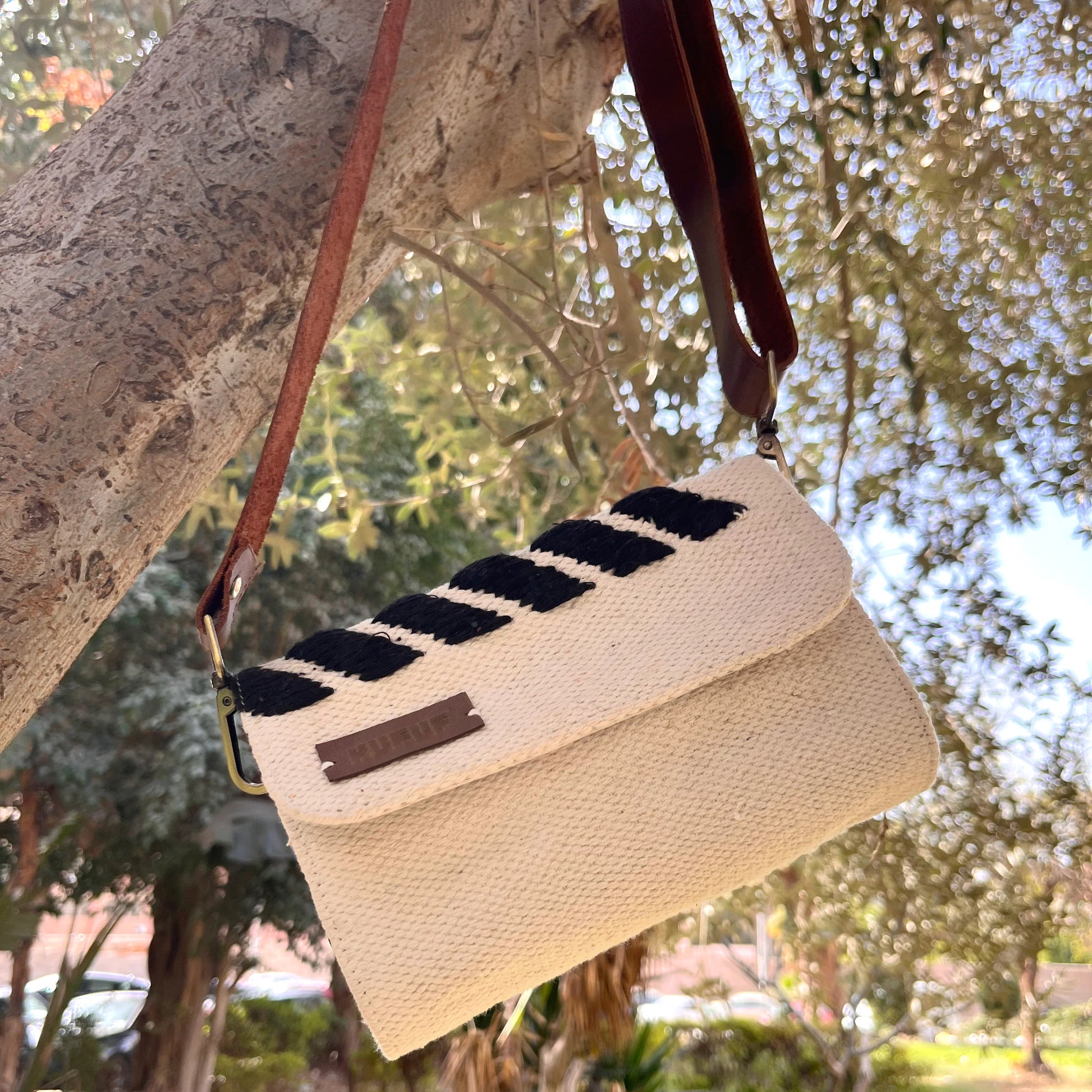 "A cream-colored woven bag with black stripes hanging from a tree branch, set against a backdrop of greenery. The bag features a brown leather strap, emphasizing its stylish design in a natural environment."