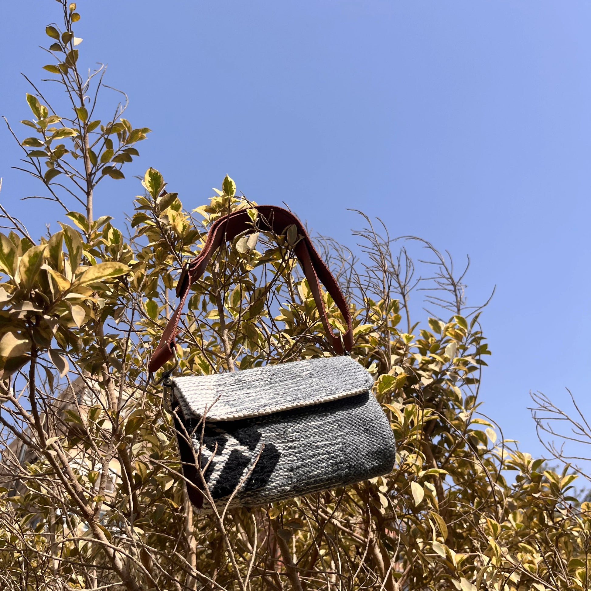  "A gray woven bag hanging from branches in a bush, set against a clear blue sky. The bag features a brown leather strap and showcases its textured design amidst natural surroundings."