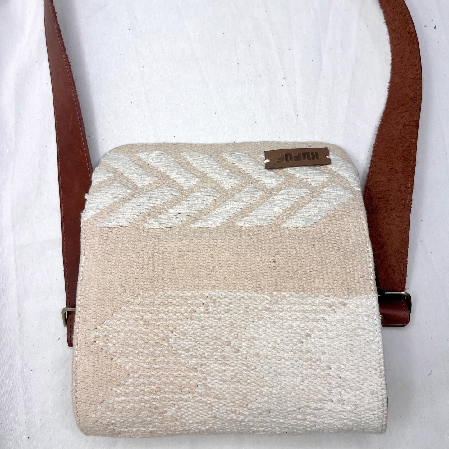 "A top-down view of a cream-colored woven bag with a white chevron design and brown leather strap, resting on a white surface. The image emphasizes the bag's clean lines and texture."