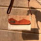  A stylish woven handbag with a striped tan and white design, featuring a brown flap and a metal label that reads "KUFUF." The bag is hanging against a textured stone wall, showcasing its unique craftsmanship.