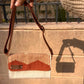 A person holding a stylish woven handbag with a striped tan and white design, featuring a brown flap and a metal label that reads "KUFUF." The bag is displayed against a textured stone wall, with a shadow cast on the surface below.