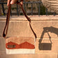 A person holding a stylish woven handbag with a striped tan and white design, featuring a brown flap and a metal label that reads "KUFUF." The bag is displayed against a textured stone wall, with a shadow cast on the surface below.