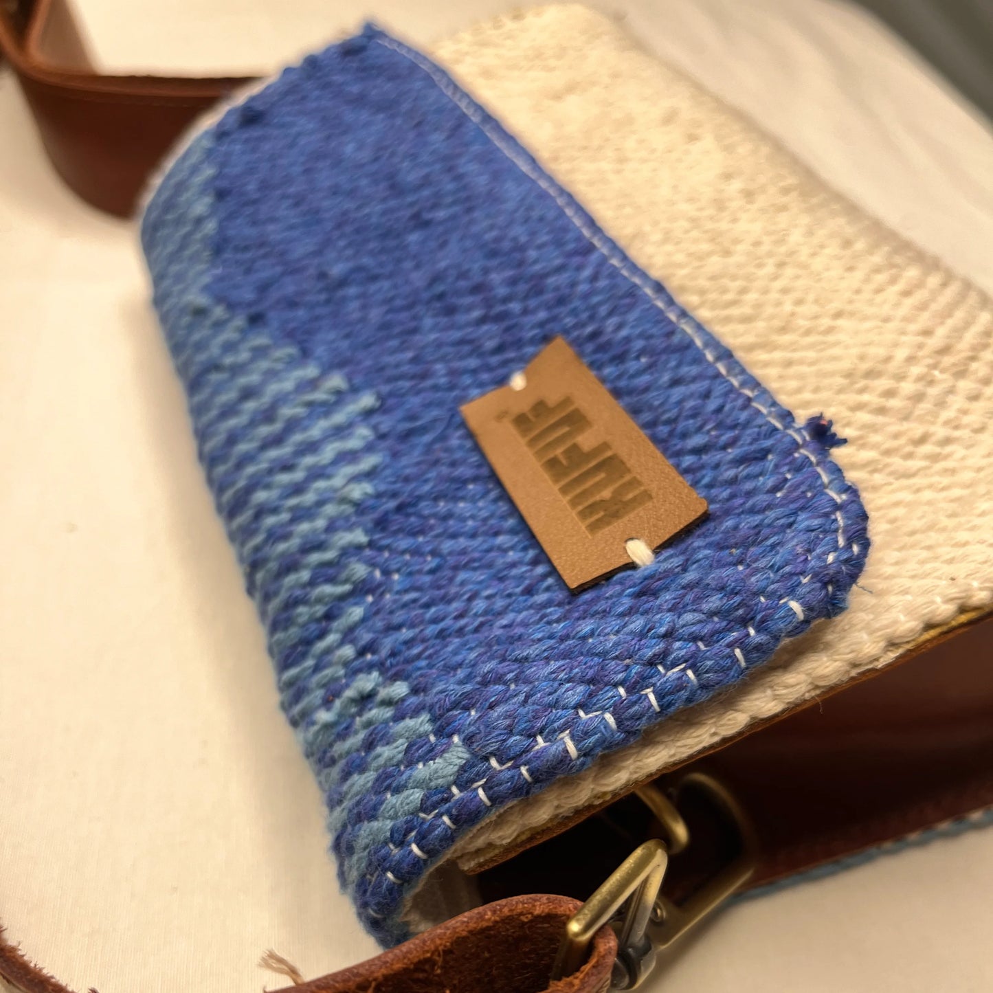 A close-up of a woven handbag shows the textured blue fabric and cream bottom. The brown leather tag with the brand name "KUFUF" is prominently displayed, highlighting the craftsmanship of the bag.