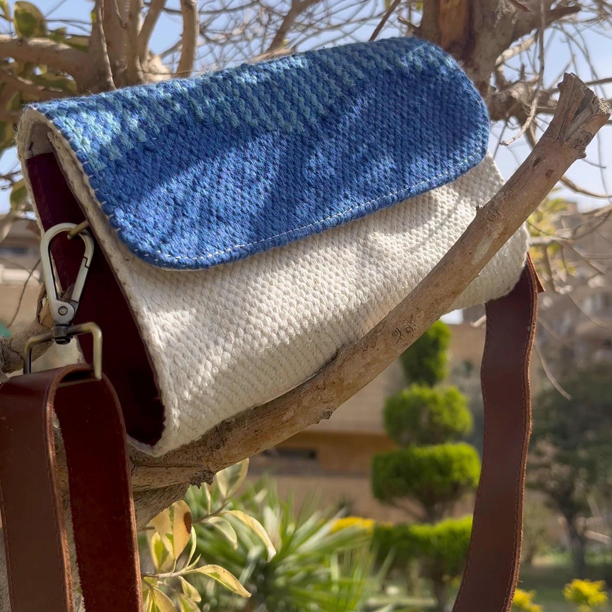 A woven handbag is showcased hanging from a branch in a tree, featuring a blue and cream design. The natural surroundings, including green foliage, create a fresh and vibrant backdrop for the bag.