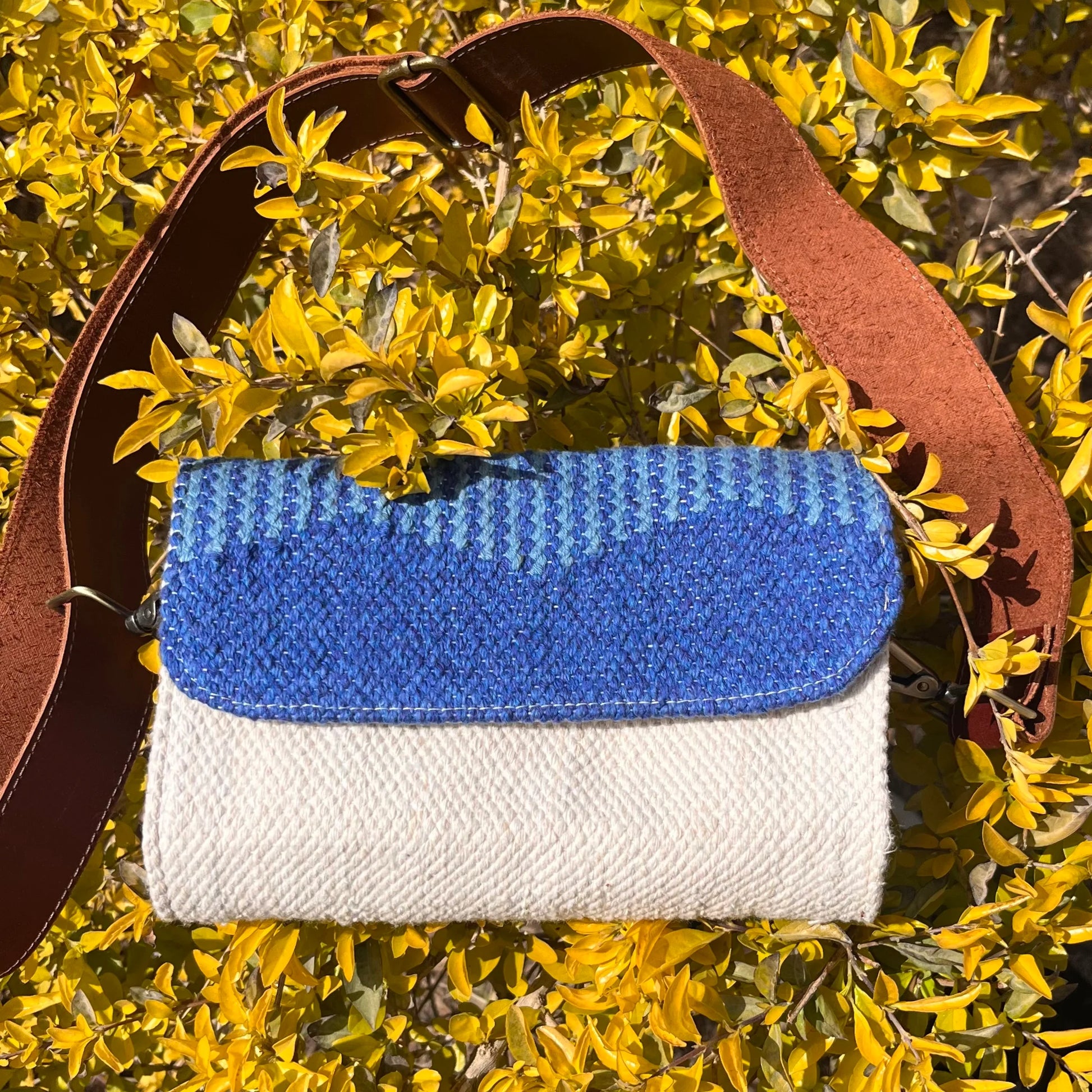 A woven handbag is placed among bright yellow foliage, showcasing its blue and cream color scheme. The brown leather strap adds a stylish touch, creating a striking contrast with the background.