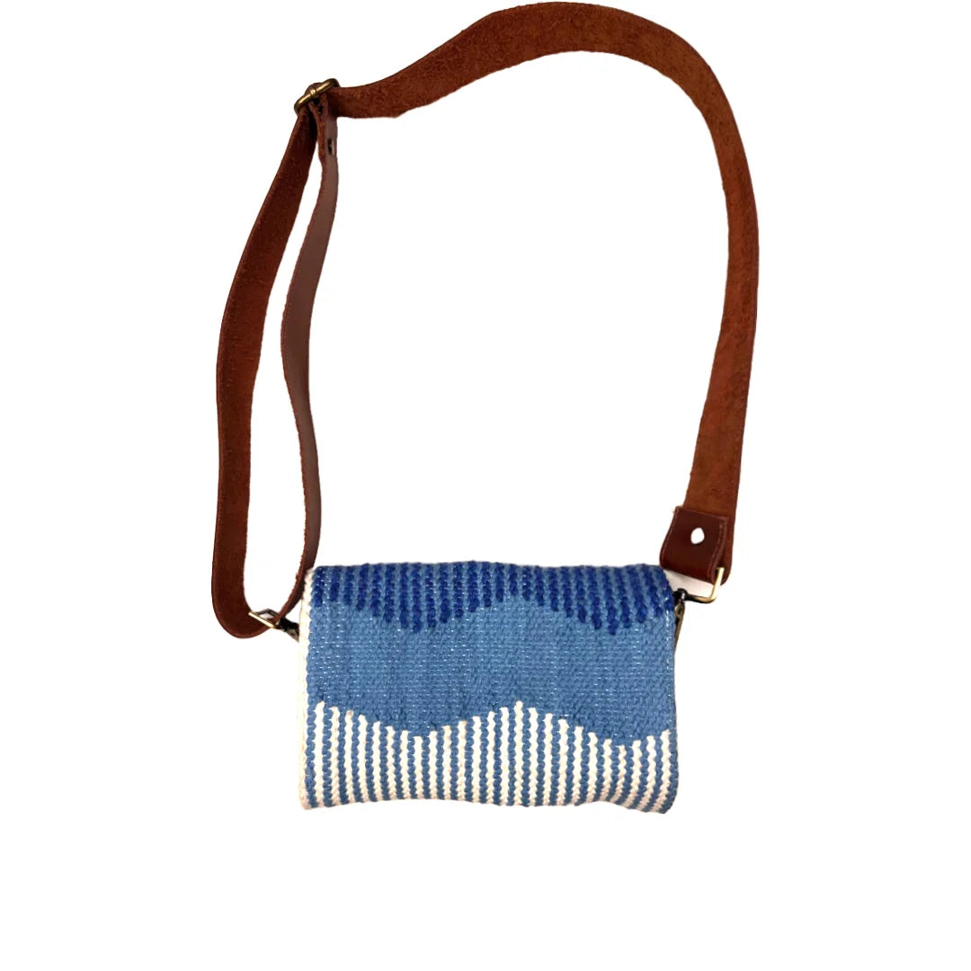 A woven handbag is presented against a white background, featuring a blue patterned top and a cream bottom. The bag has a brown leather strap and a brand tag, showcasing its modern and chic design.