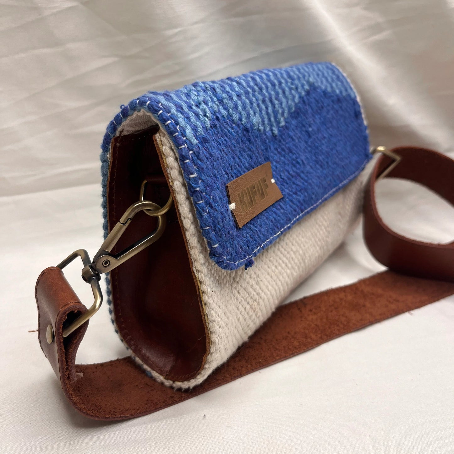 A close-up of a woven handbag highlights its side view, showcasing the textured blue fabric and cream bottom. The brown leather strap and a small brand tag labeled "KUFUF" enhance the bag's overall aesthetic.